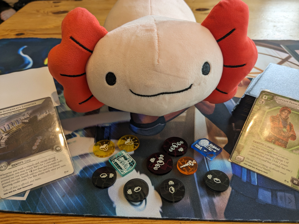 a cute axolotl plushie sits on a netrunner playmat, accompanied by my decks (with the identity cards Ob and Padma facing the viewer) and a collection of lovely acrylic netrunner tokens