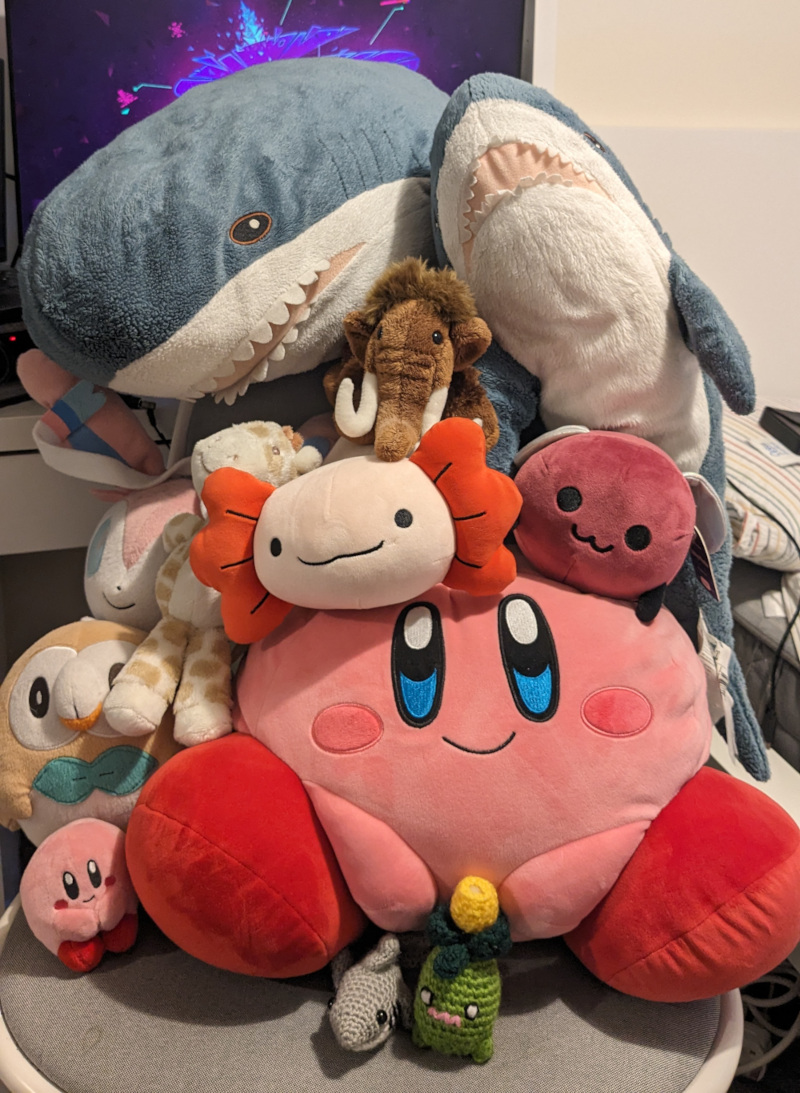 a stack of plushies, featuring a sylveon, rowlett, tiny kirby, big kirby, smoliv, tiny shark, giraffe, medium blahaj, big blahaj, axi the axolotl, a mammoth and eggbug!